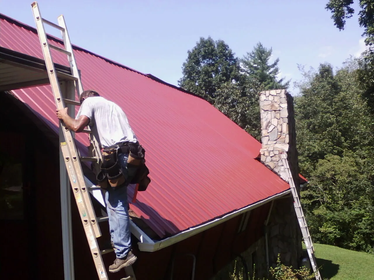 Roofer on a ladder beside a red metal roof and stone chimney — Soffit & Fascia Repair by Roofing Brothers in Royal